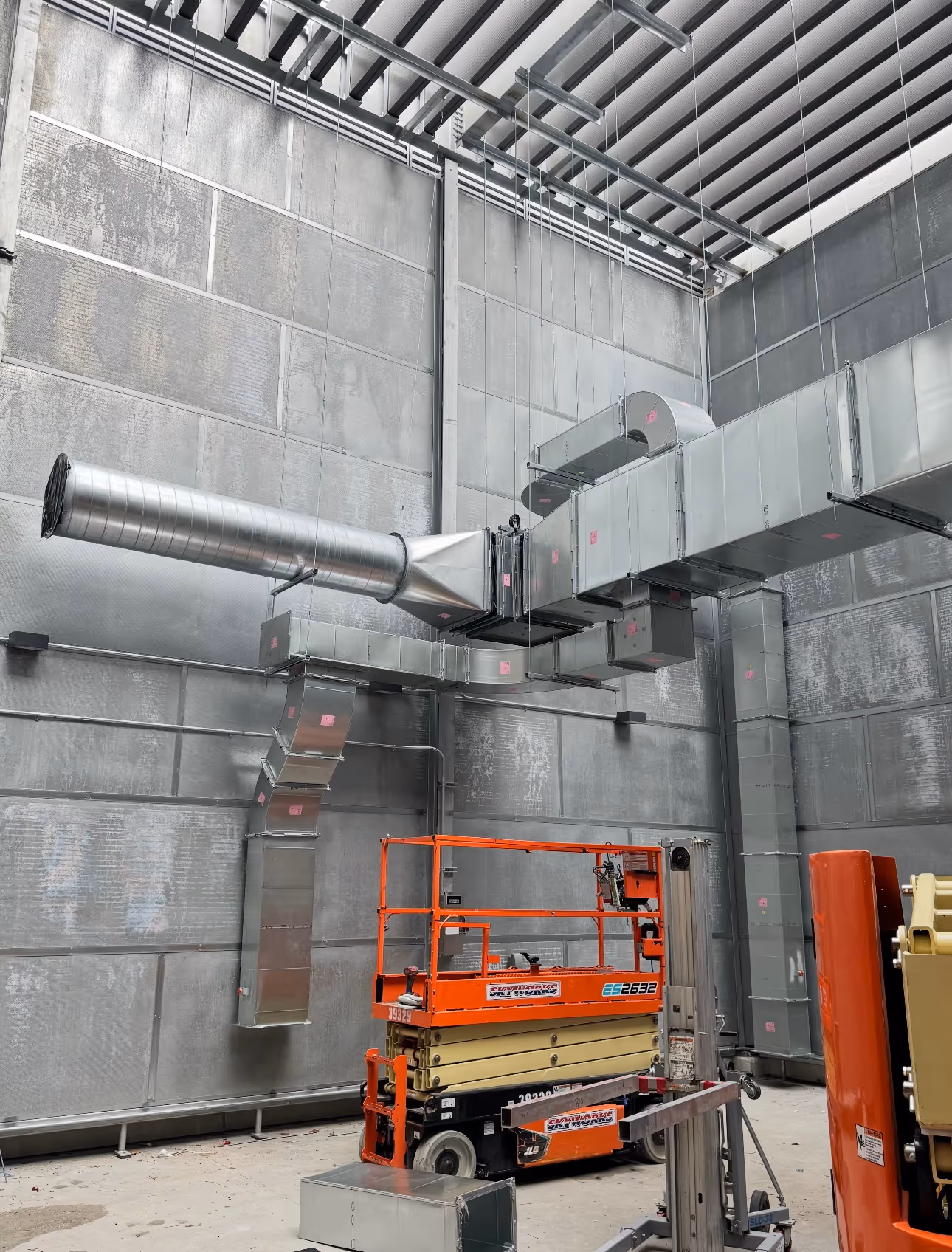 Industrial ventilation ducts installed on a warehouse wall with a scissor lift in front.