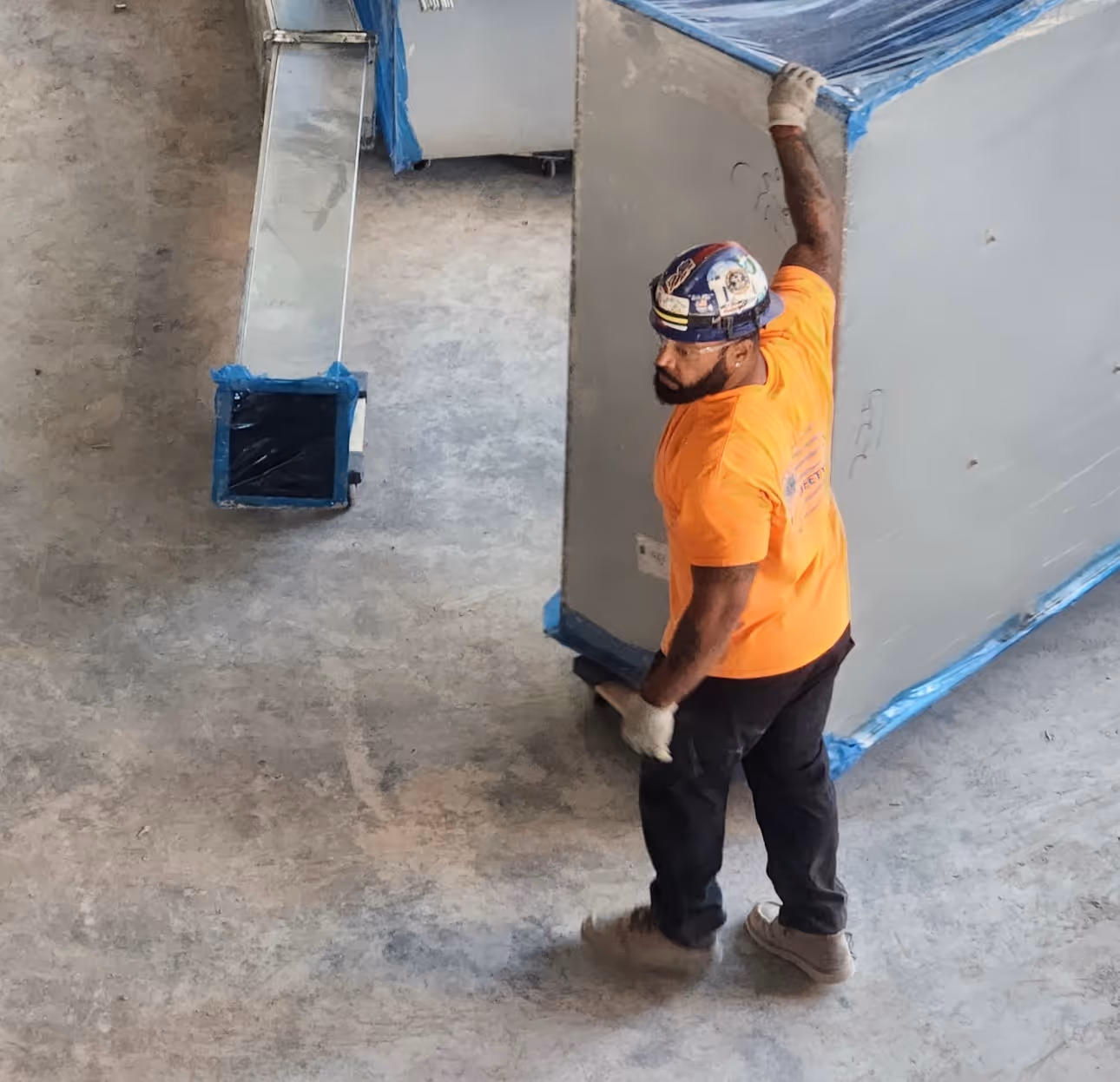 Construction worker in orange shirt and hard hat moving large wrapped metal duct.