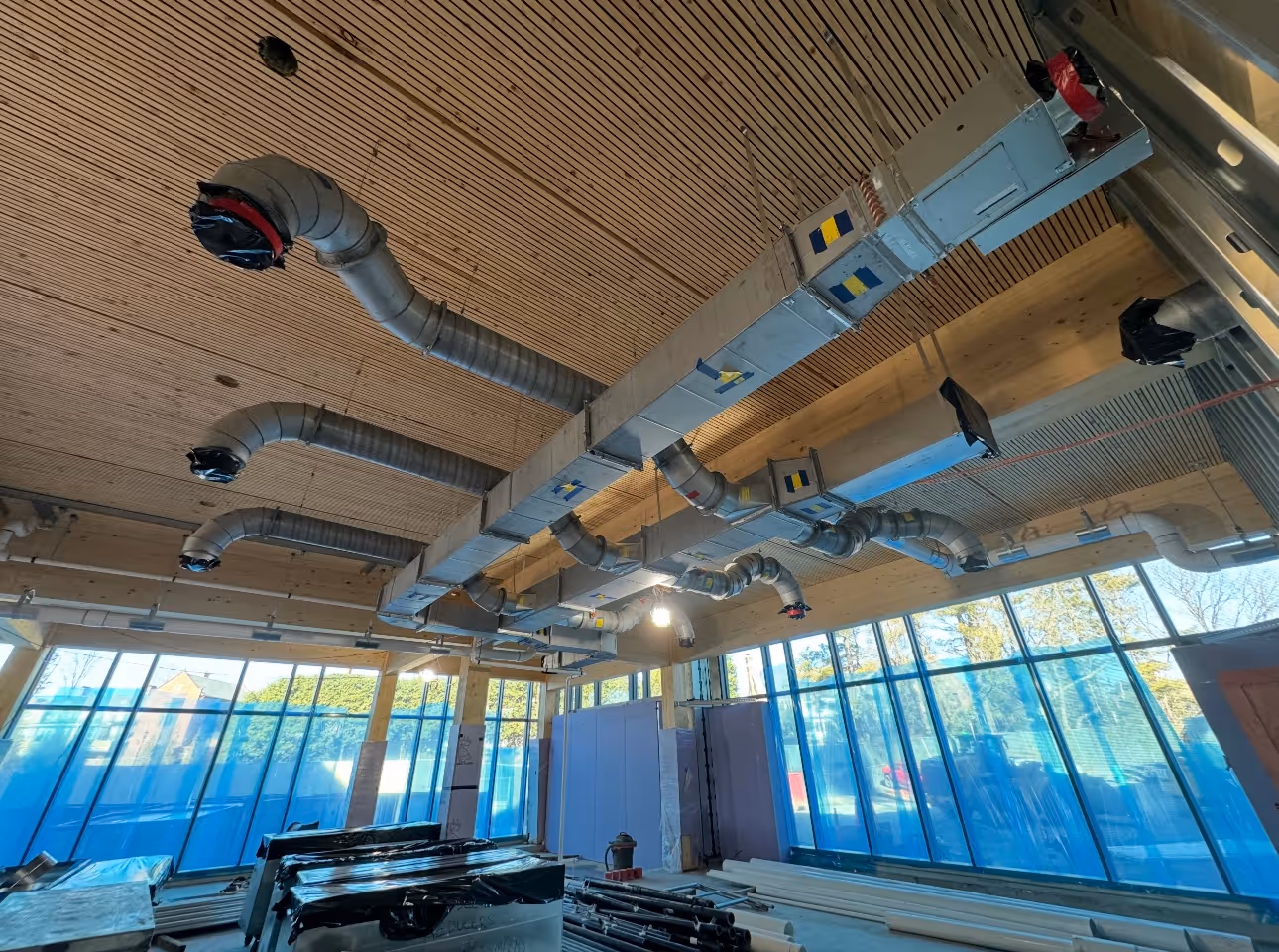 Ceiling with complex HVAC ductwork in a room under construction with large windows covered in blue plastic.