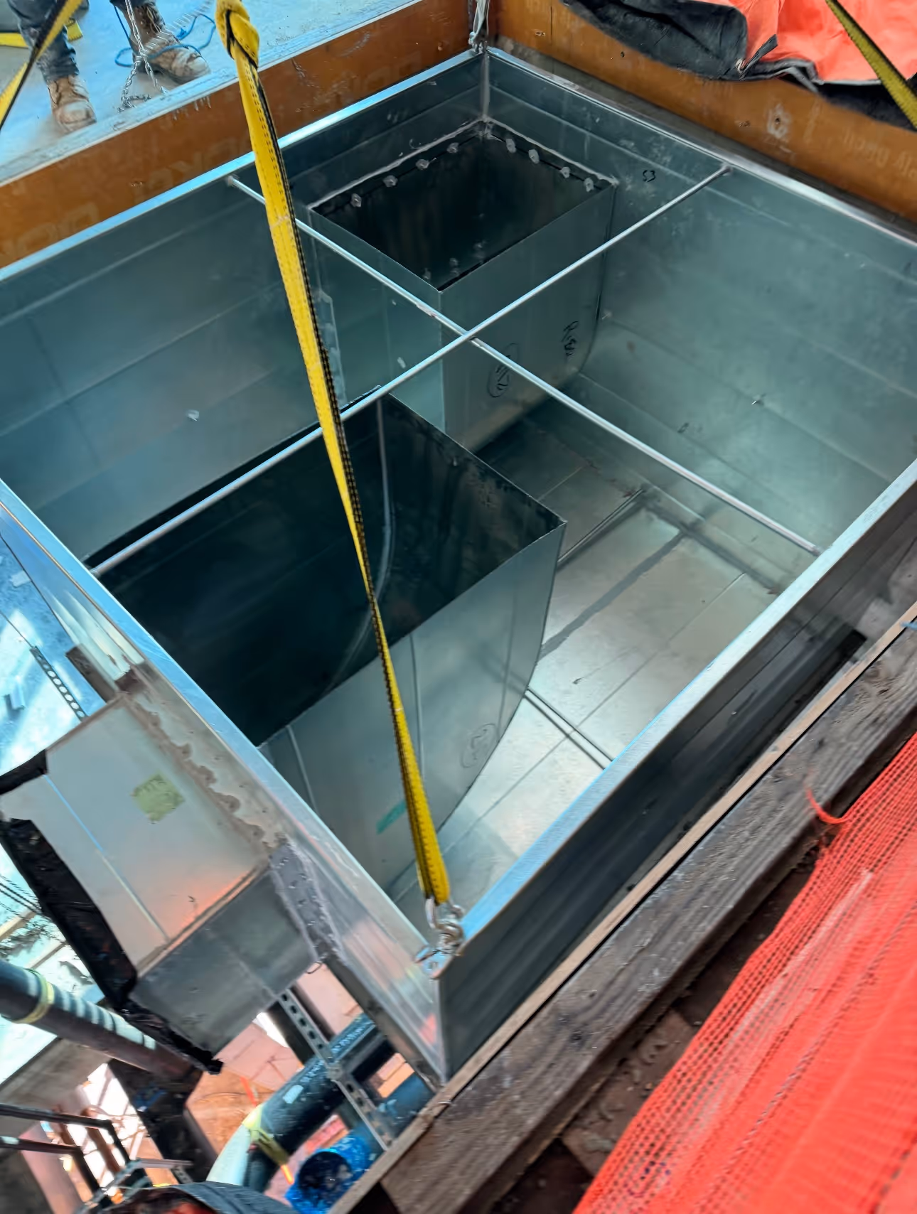 Metal ductwork section suspended by yellow strap at a construction site with workers visible.