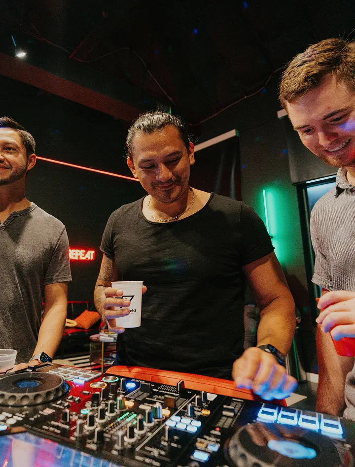Three men smiling while one of them mixes music on a DJ controller at a party.