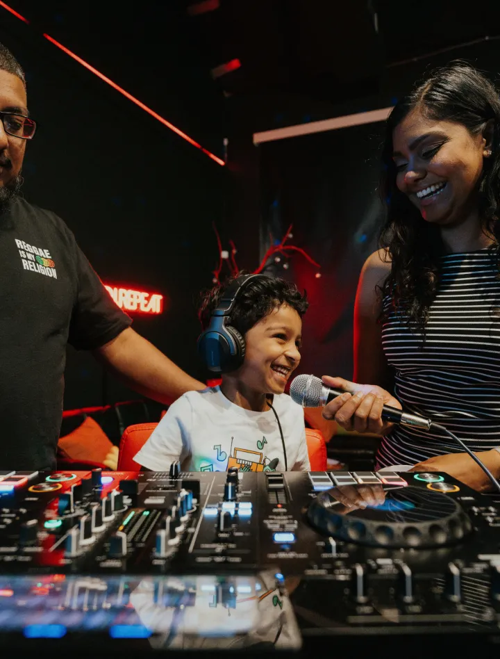A child wearing headphones smiles and sings into a microphone while adults stand beside him at a DJ setup.