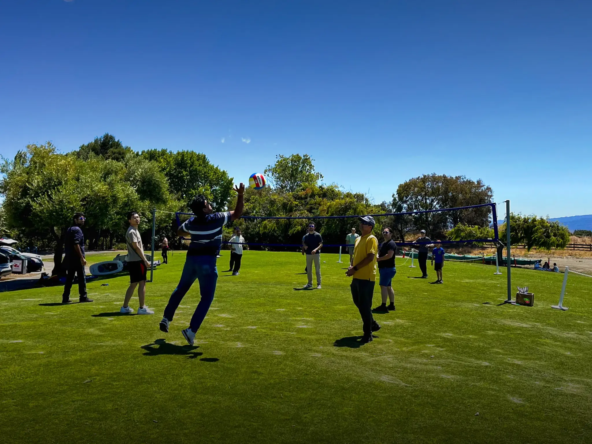 Group of people playing volleyball on a grassy field under clear blue sky.