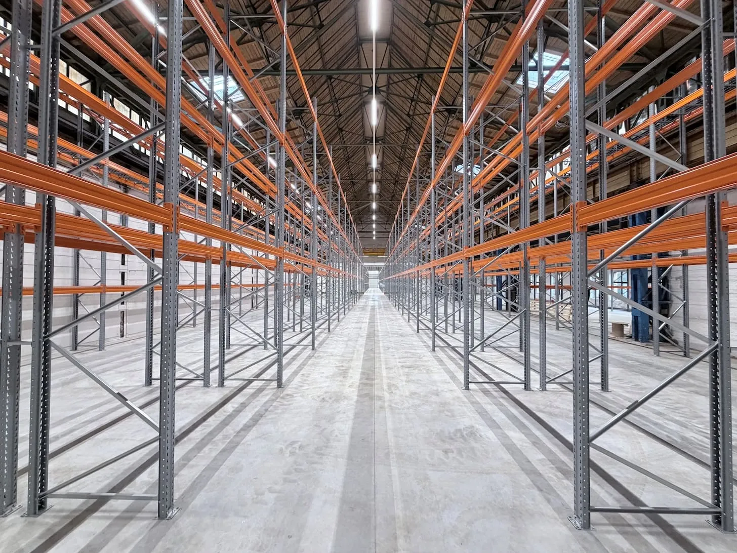Leerer Lagerraum mit hohen Metallregalen und orangefarbenen Trägern unter einem hohen, beleuchteten Dach.