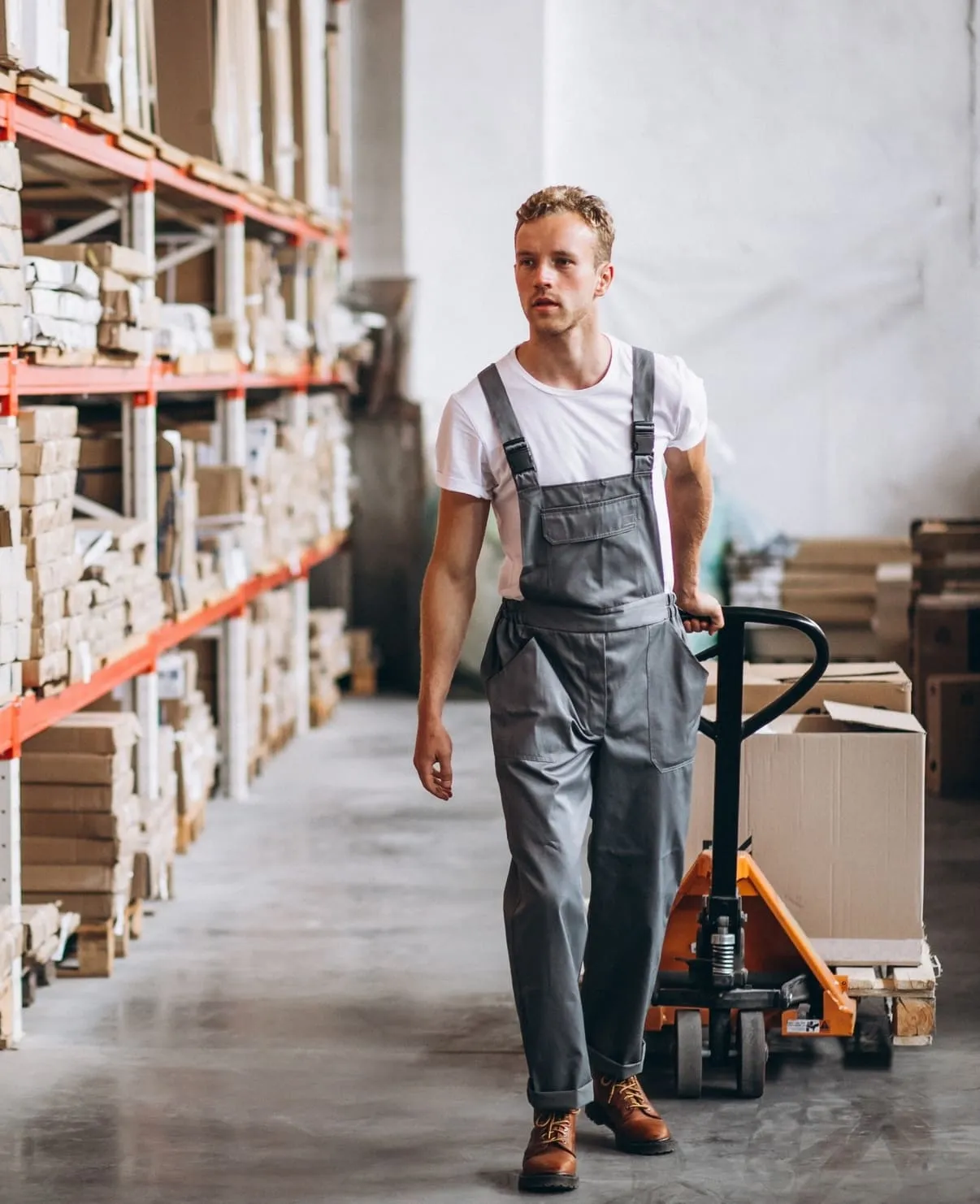 Ein Lagerarbeiter in grauer Arbeitskleidung zieht eine Palette mit Kartons auf einem Hubwagen in einem Lager.