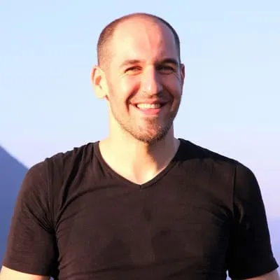 Lächelnder Mann mit kurz geschorenem Haar und schwarzem T-Shirt vor klarem Himmel.