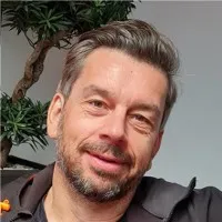 Porträt eines lächelnden Mannes mit kurzen braunen Haaren und Bart vor einer Pflanze und grauem Hintergrund.