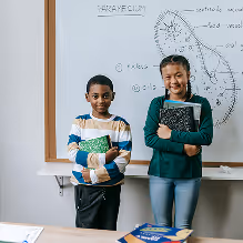 Two smiling students standing in front of a whiteboard with scientific diagrams, one holding a notebook.