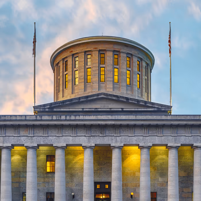 Evening view of a classical government building with tall columns, lit windows, and two American flags.