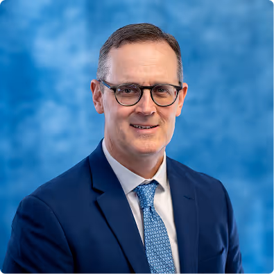 Middle-aged man wearing glasses, a navy blue suit, white shirt, and patterned tie against a blue blurred background.