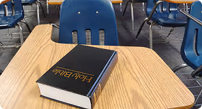 Black Holy Bible with gold lettering lying on a wooden school desk in a classroom.