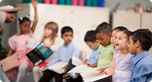 Diverse group of young children sitting in a classroom, reading books and raising hands.