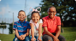 Three smiling children sitting on grass outdoors on a sunny day.