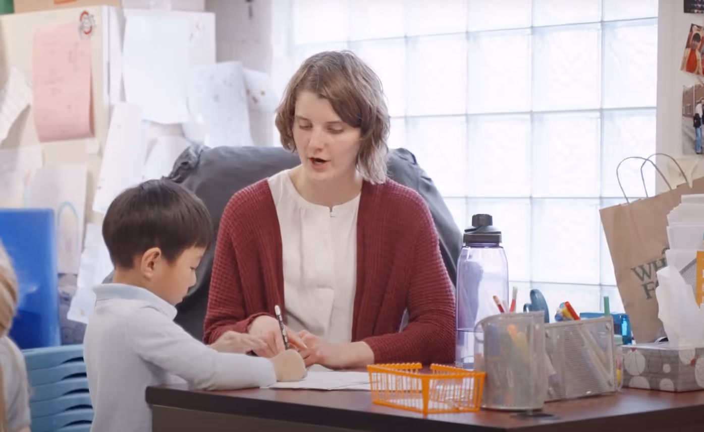 Teacher in a red cardigan assisting a young boy who is writing on paper at a classroom table.