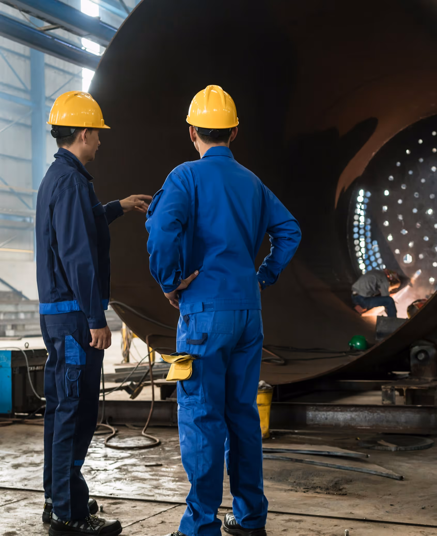Two factory workers wearing yellow helmets and blue uniforms inspecting a large cylindrical metal structure while a third worker welds inside it.