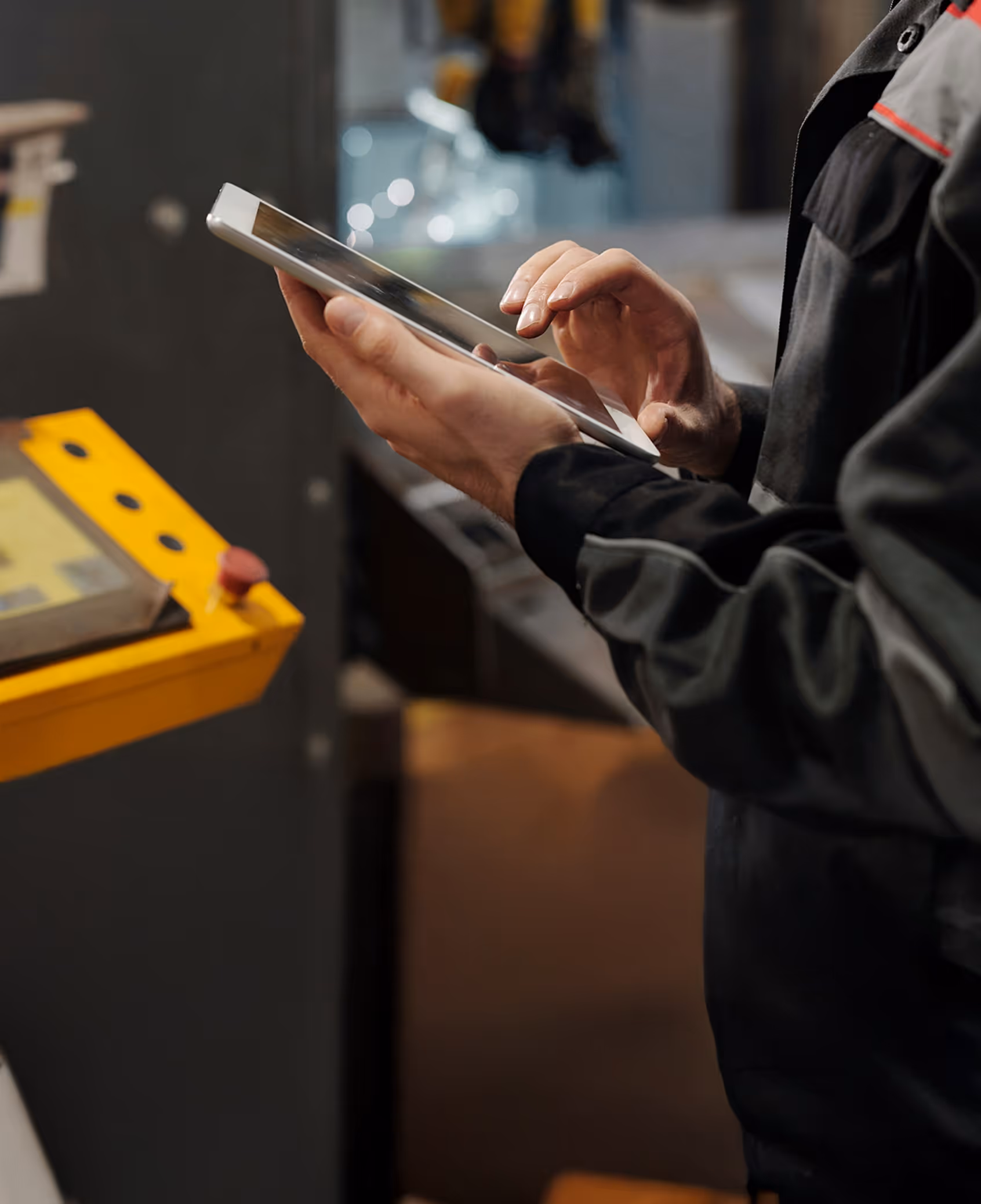 Person in work attire using a tablet in an industrial setting near yellow control panel.
