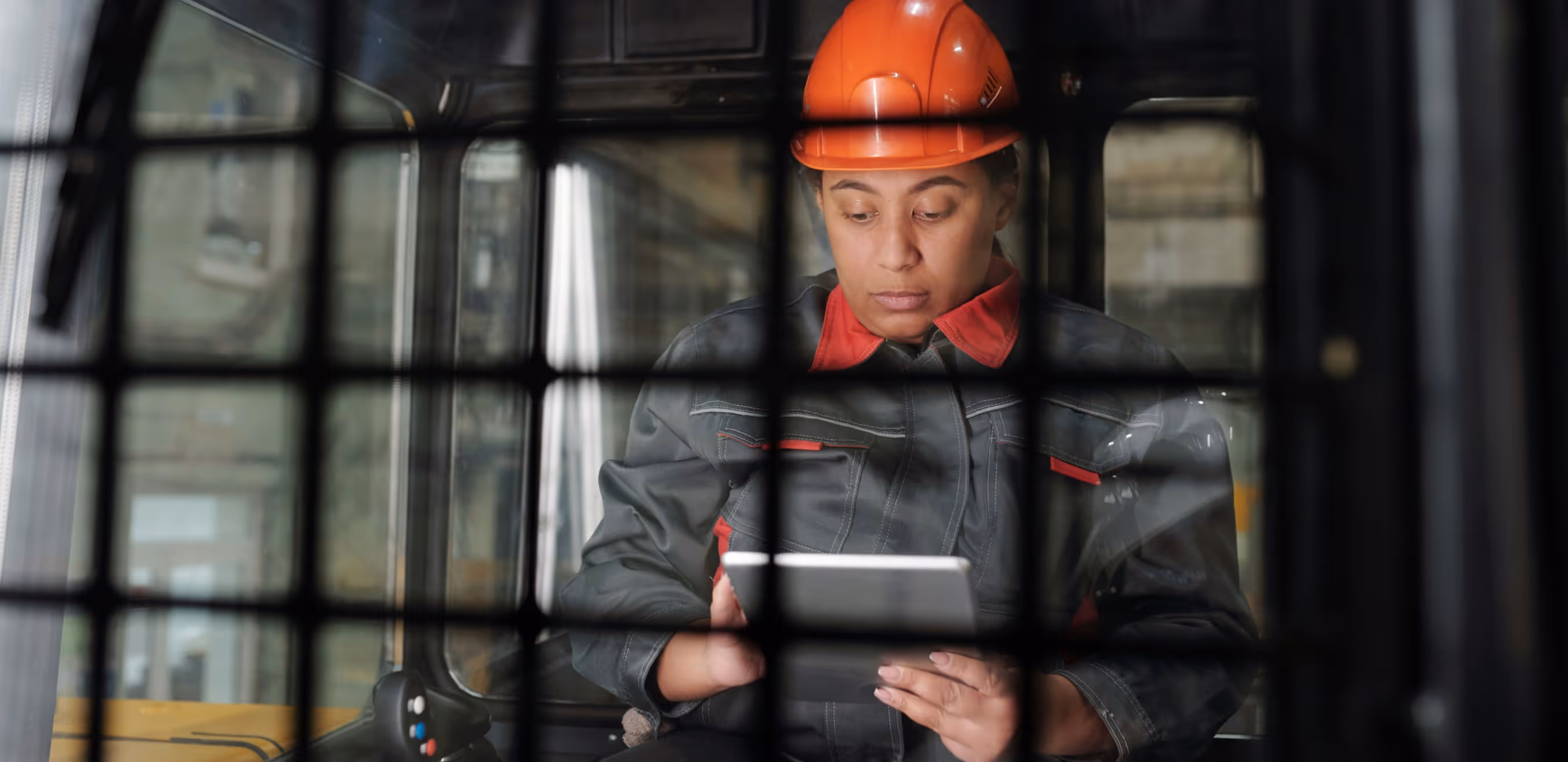 Worker wearing an orange hard hat and dark work uniform looking at a tablet inside a vehicle with a protective grid in the foreground.