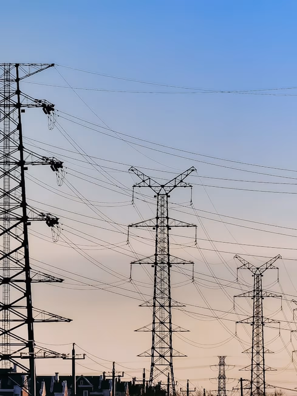 Multiple electrical transmission towers with power lines stretching across a clear sky above residential rooftops.