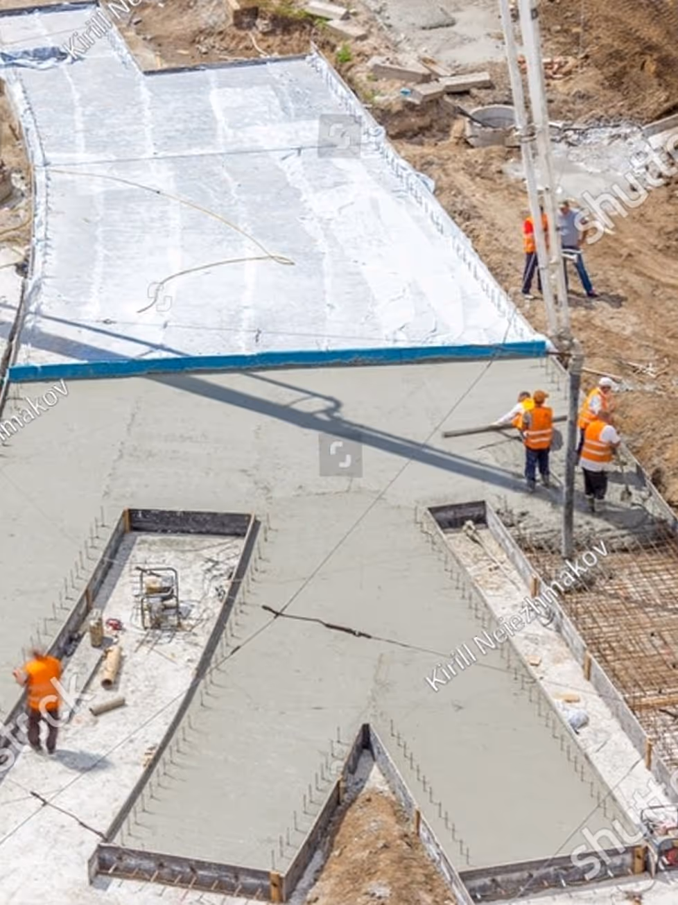 Construction workers pouring and leveling concrete on a large cross-shaped foundation slab at a building site.