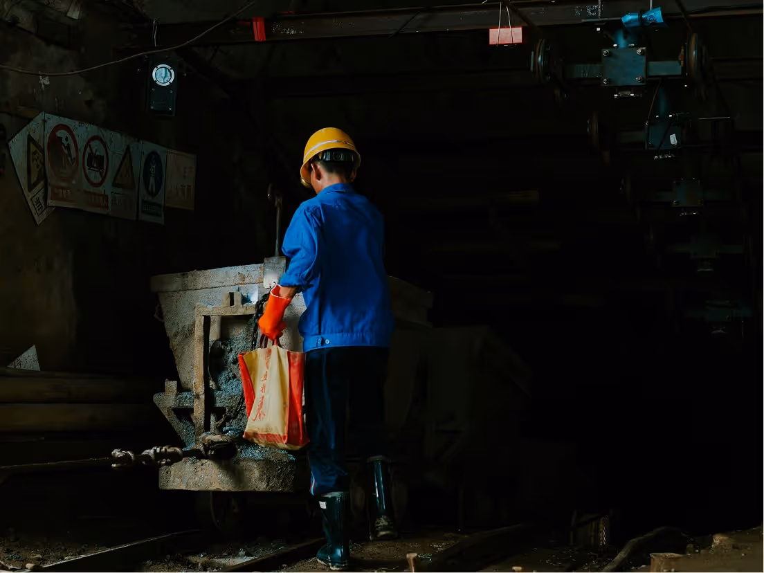Worker in blue uniform, yellow helmet, and red gloves operating machinery in a dark industrial setting.