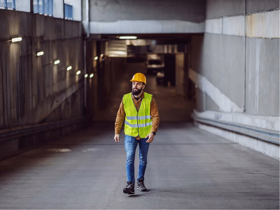 Construction worker wearing a yellow hard hat and reflective vest walking in a concrete tunnel.