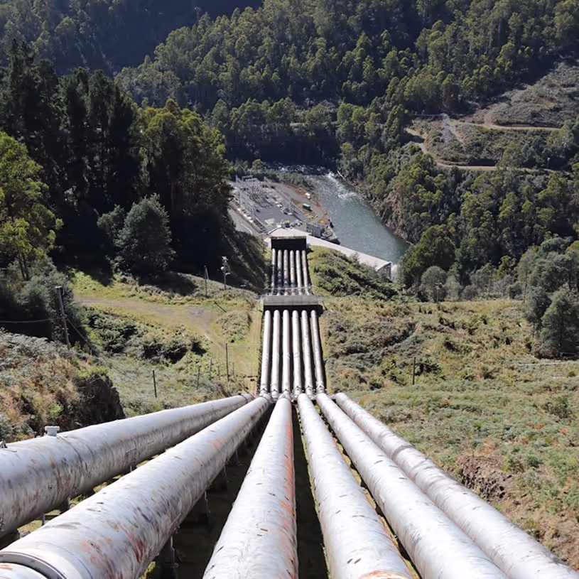 Large industrial water pipes descending a steep hill toward a river surrounded by forested mountains.