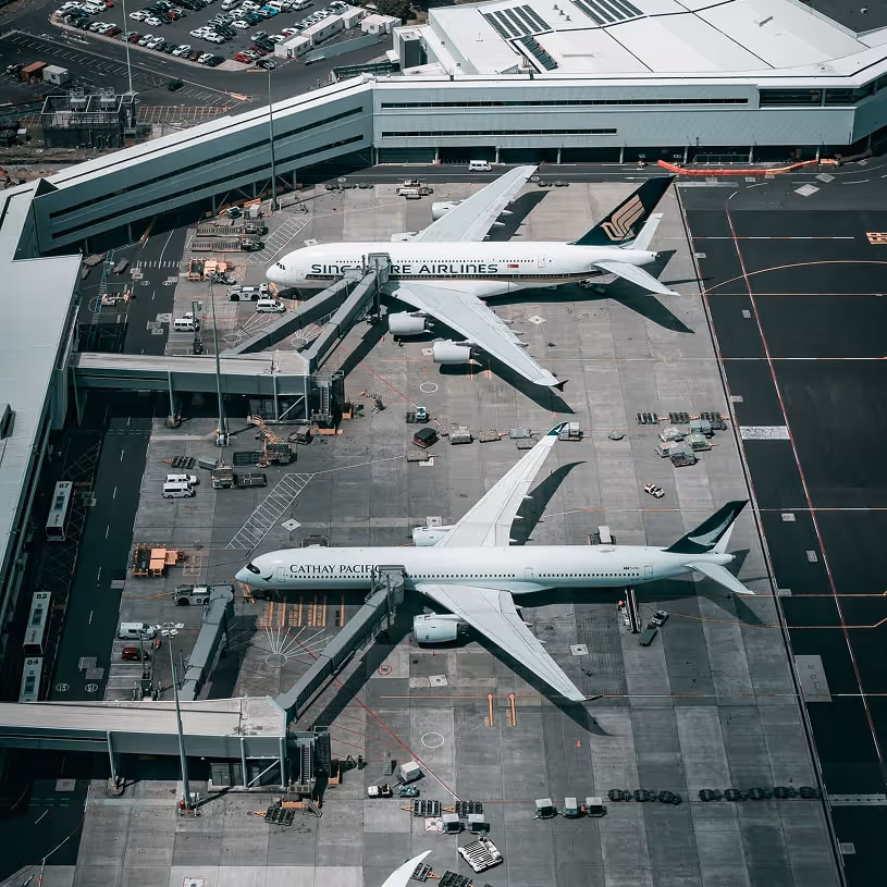 Aerial view of two airplanes, one Singapore Airlines and one Cathay Pacific, parked at airport gates with jet bridges connected.