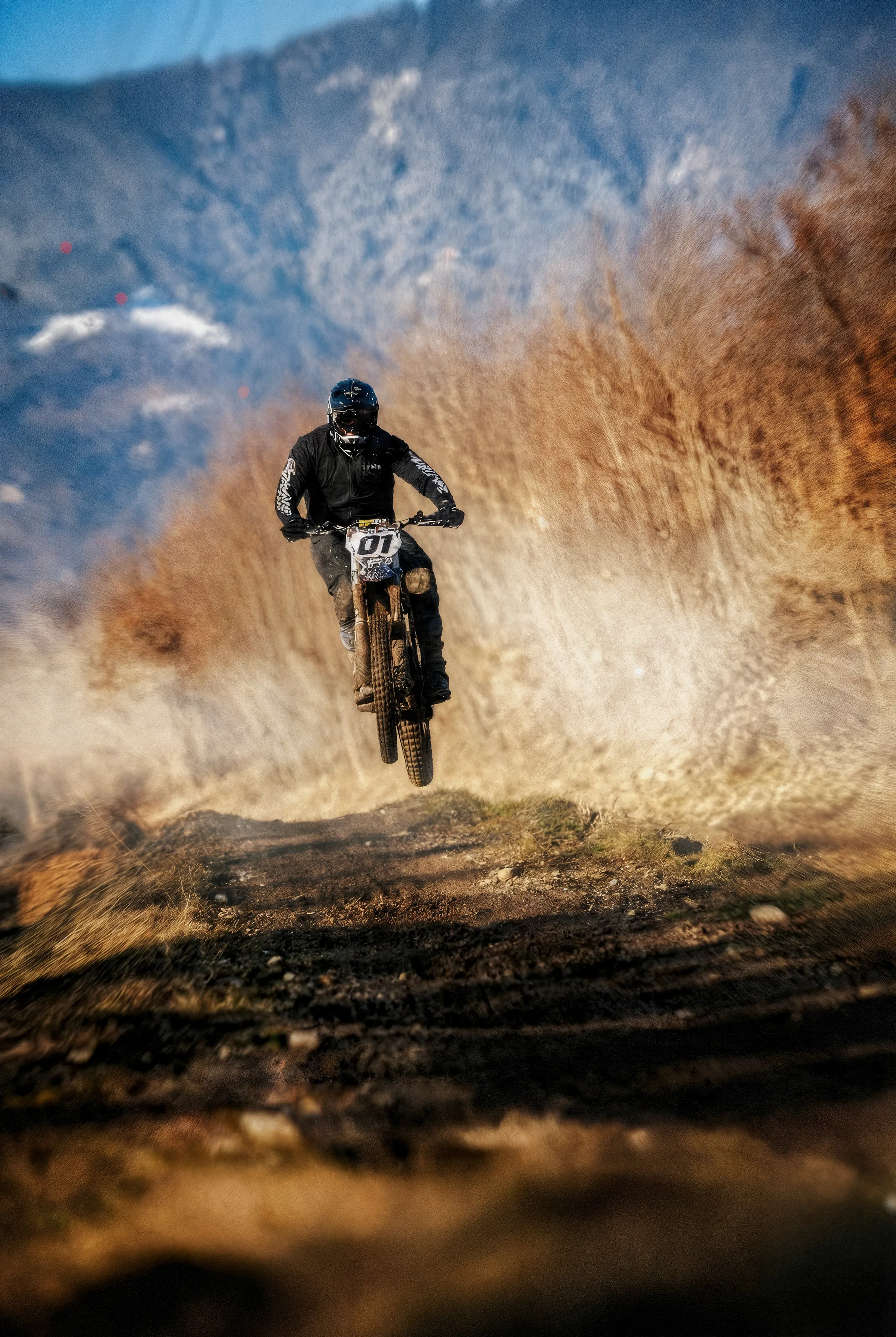 Motorcyclist in black gear airborne on a dirt trail with dust clouds and mountains in the background.