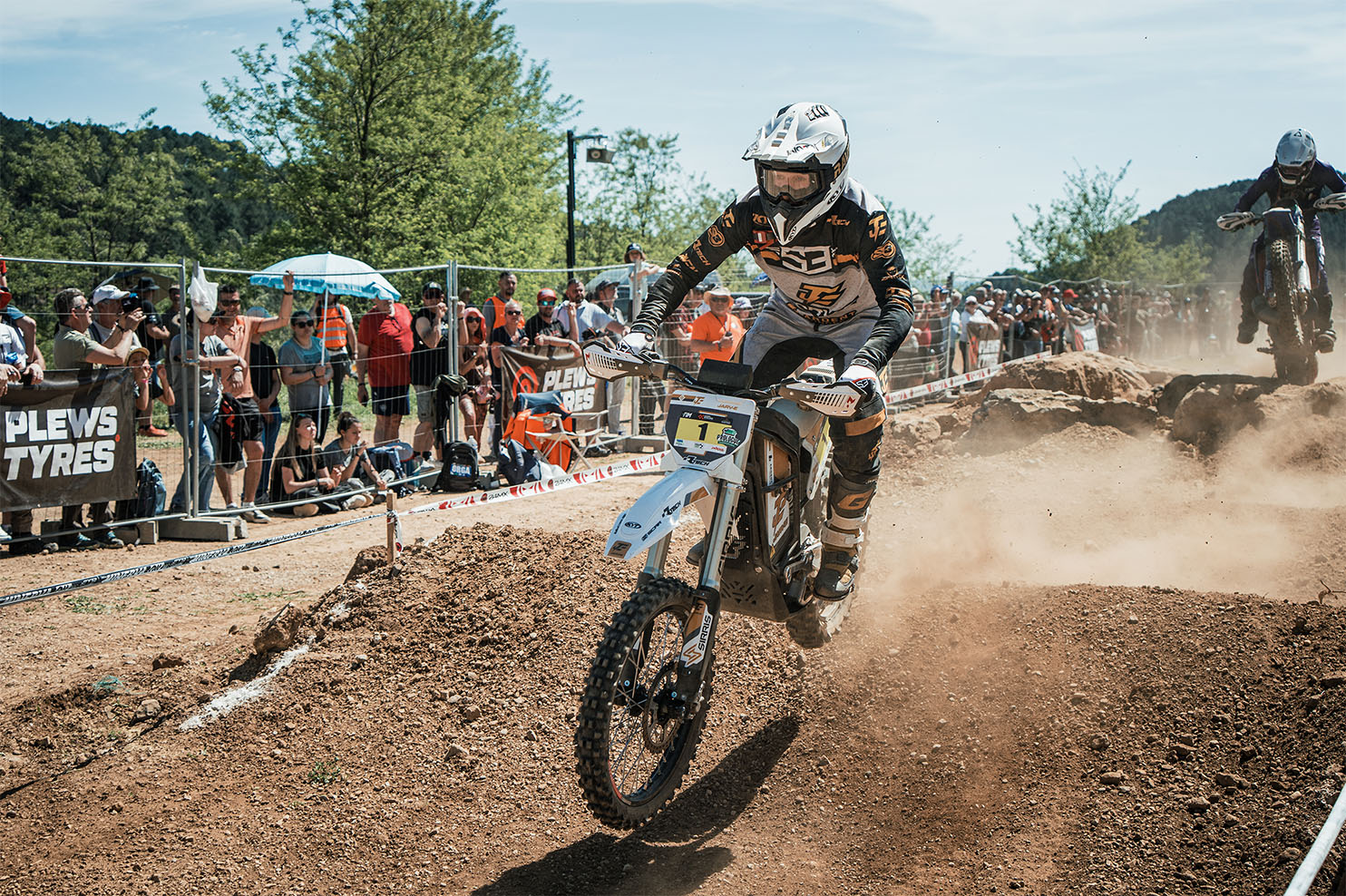 Motorcyclist riding an off-road bike on a grassy mountain slope under a partly cloudy blue sky.
