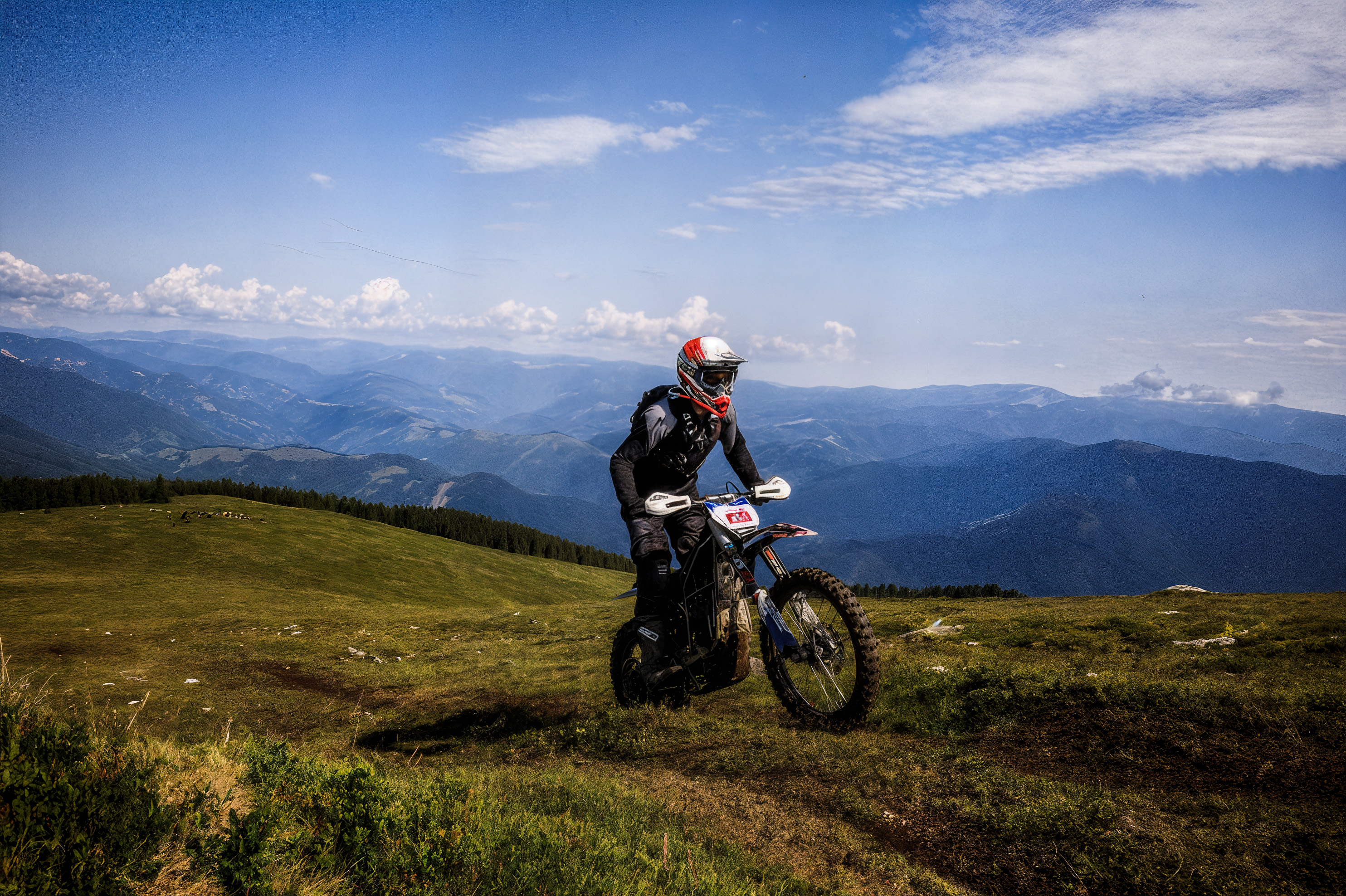 Motorcyclist riding an off-road bike on a grassy mountain slope under a partly cloudy blue sky.