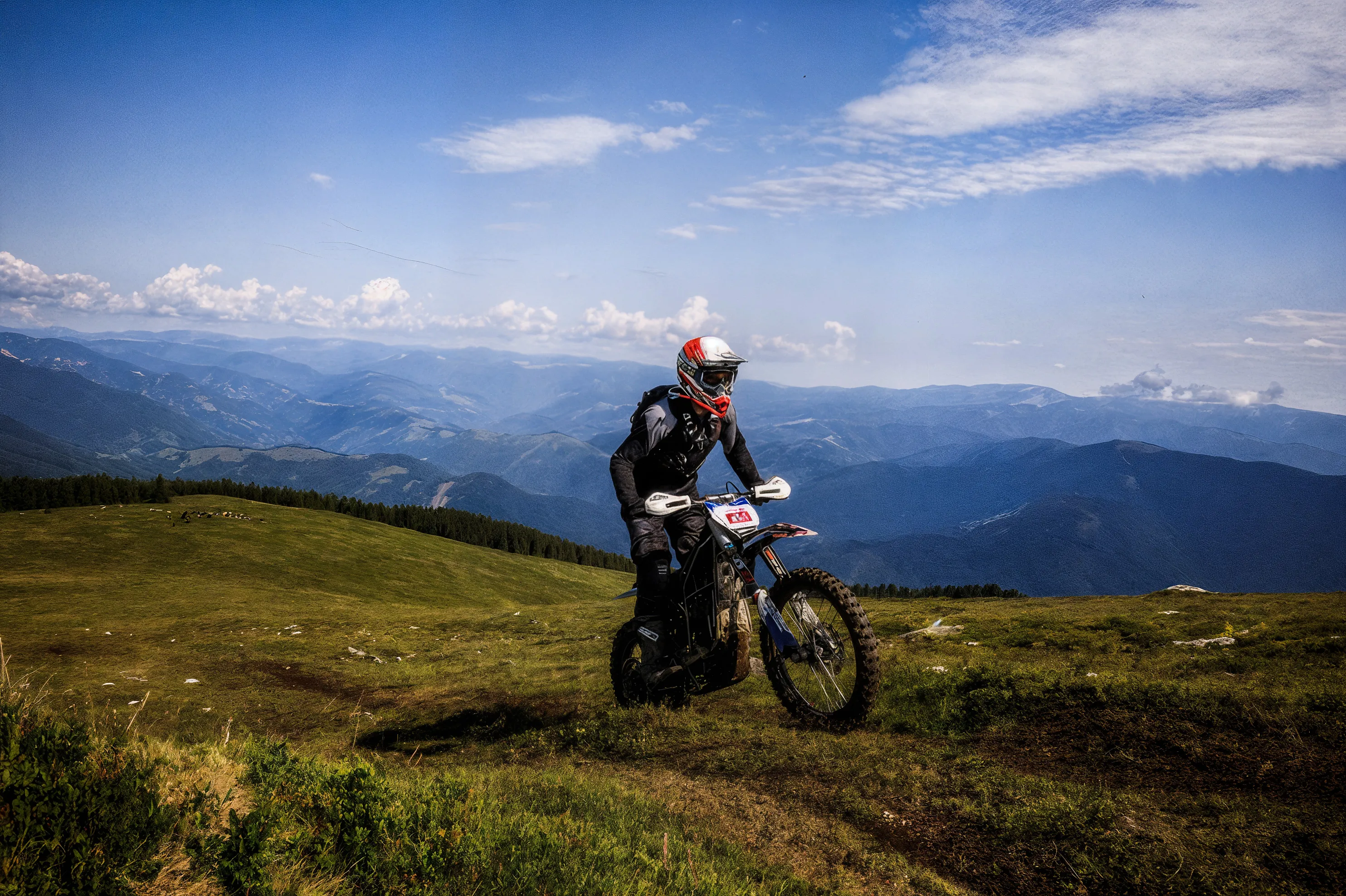 Motorcyclist riding an off-road bike on a grassy mountain slope under a partly cloudy blue sky.