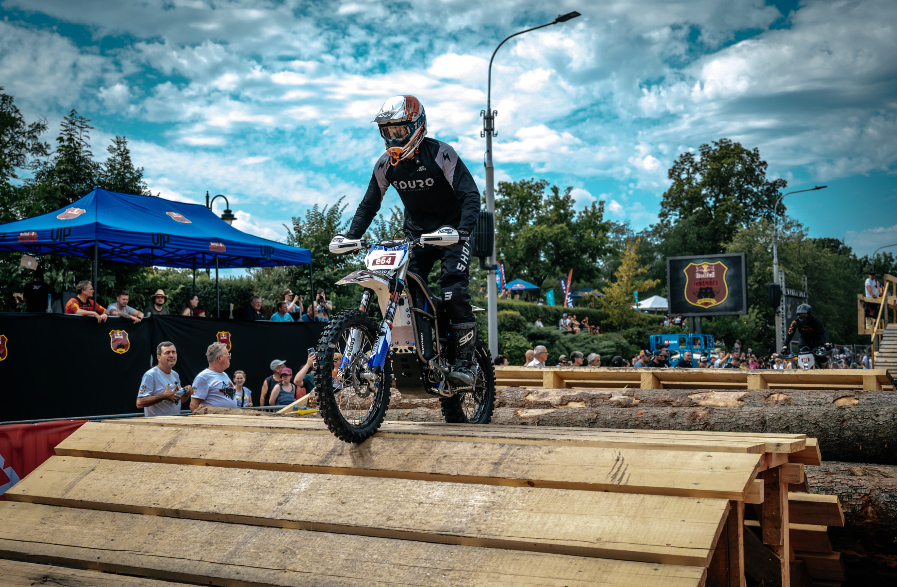 Motorcyclist wearing black gear rides an off-road bike over wooden planks during a daytime outdoor event with spectators and tents in the background.