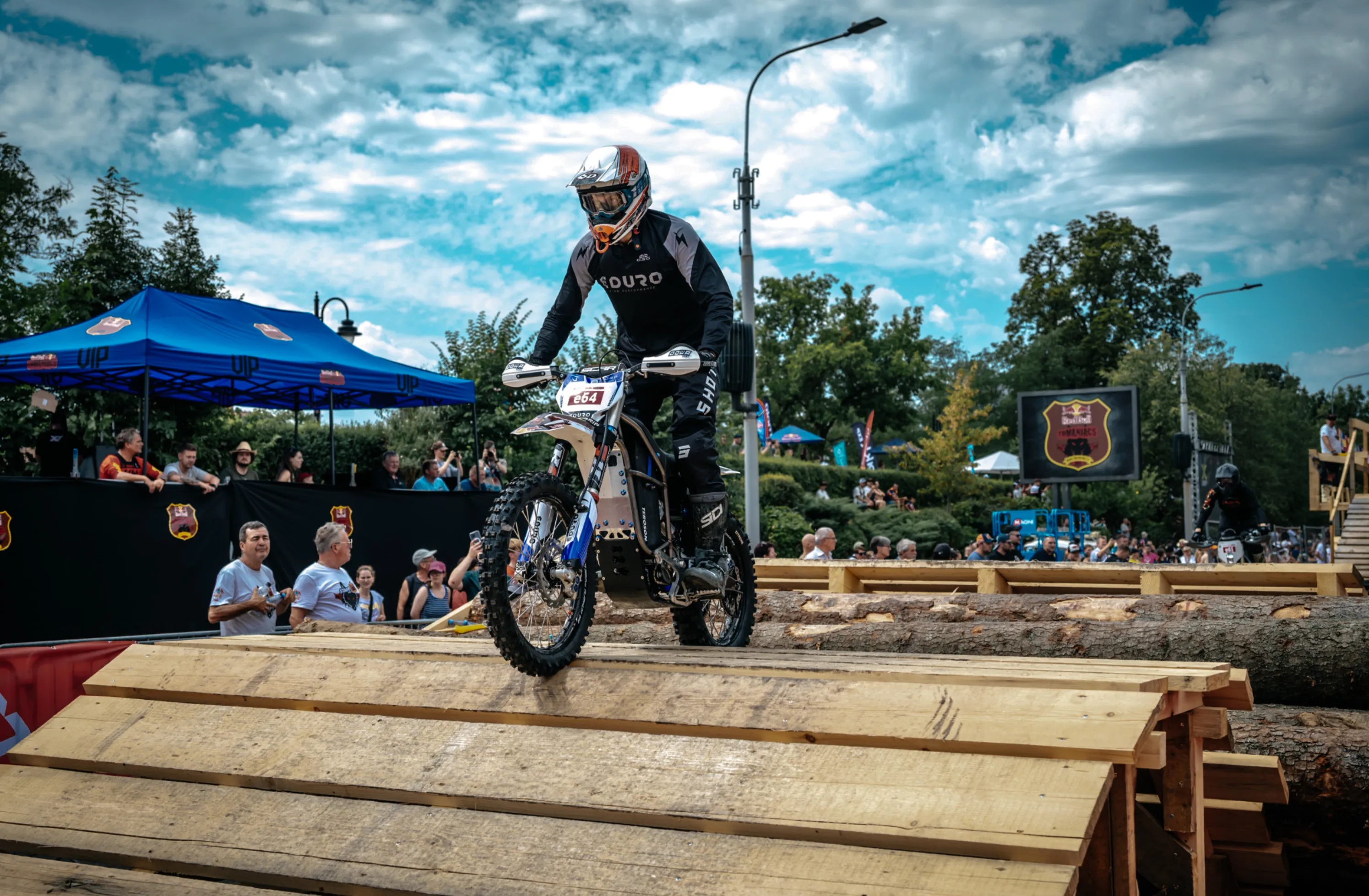 Motorcyclist wearing black gear rides an off-road bike over wooden planks during a daytime outdoor event with spectators and tents in the background.