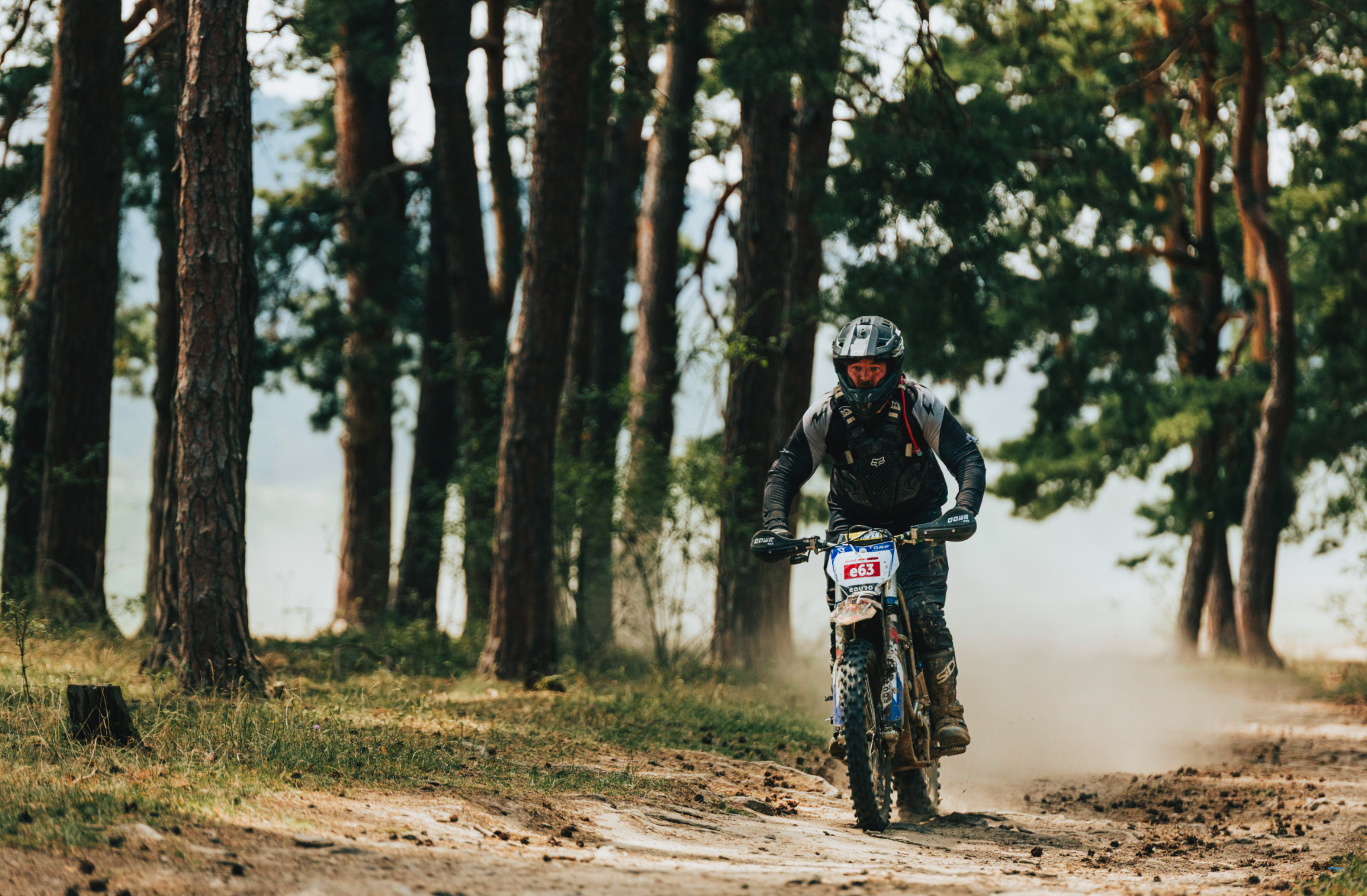 Motorcyclist wearing protective gear riding a dirt bike on a forest trail, kicking up dust.