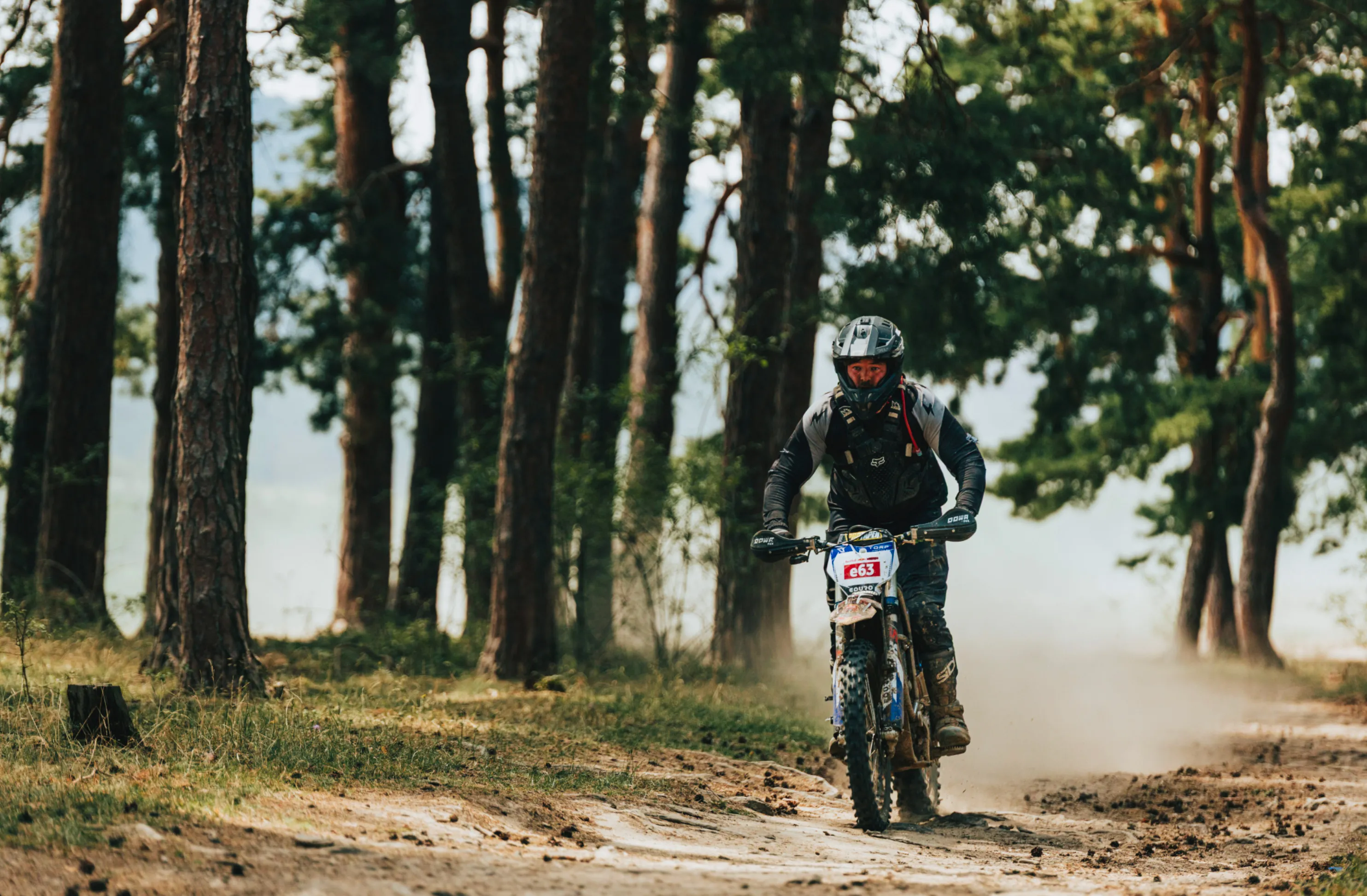 Motorcyclist wearing protective gear riding a dirt bike on a forest trail, kicking up dust.