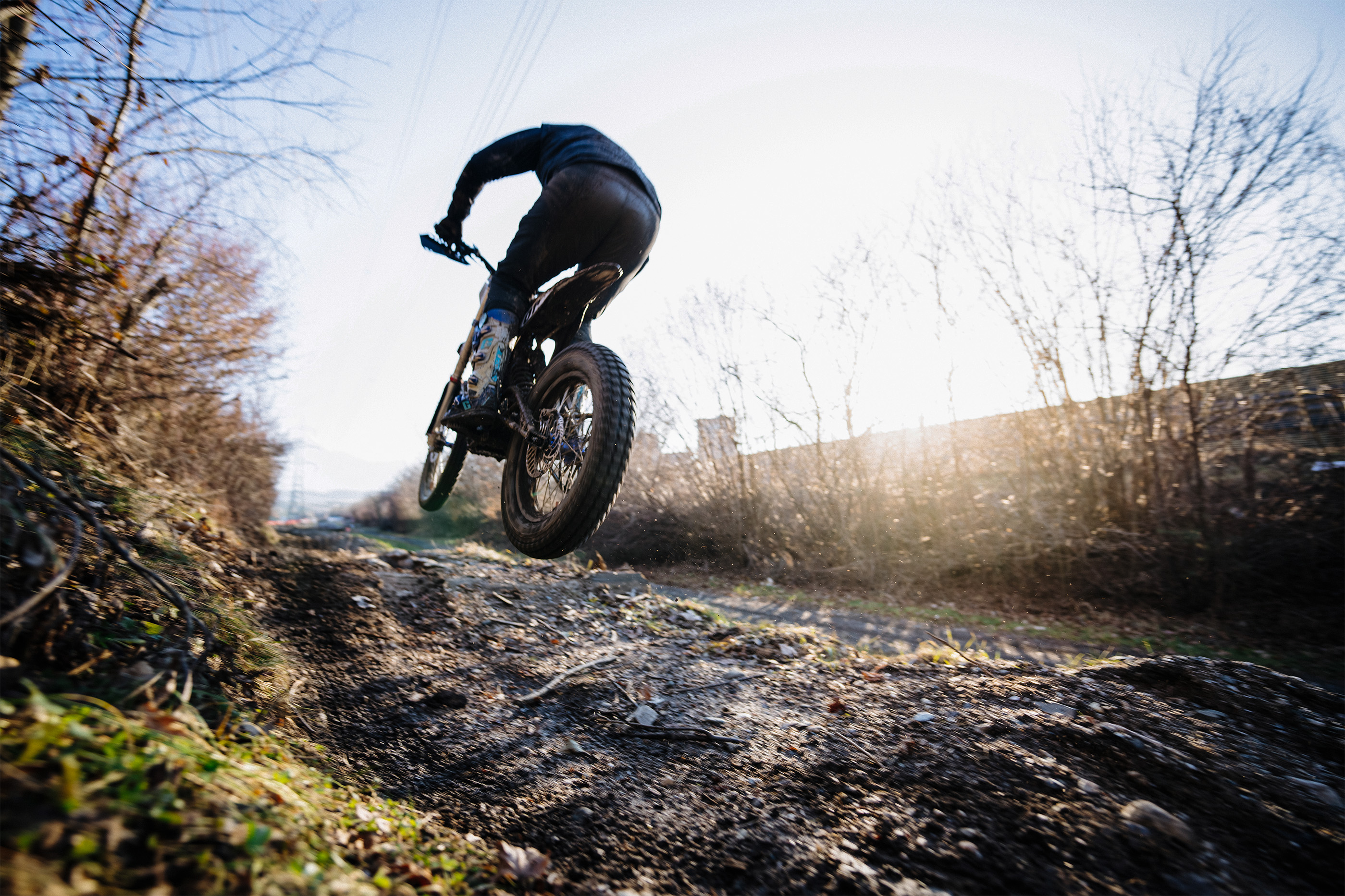 Person wearing protective gear riding a dirt bike jumping off a rough dirt trail in a wooded area.