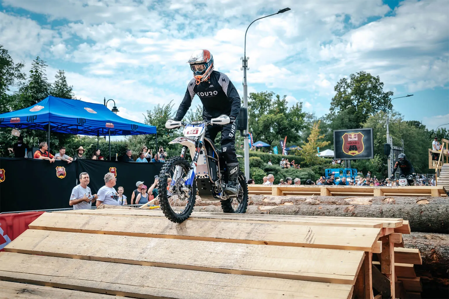 Motorcycle rider in black gear and helmet navigating wooden ramps during an outdoor event with spectators.