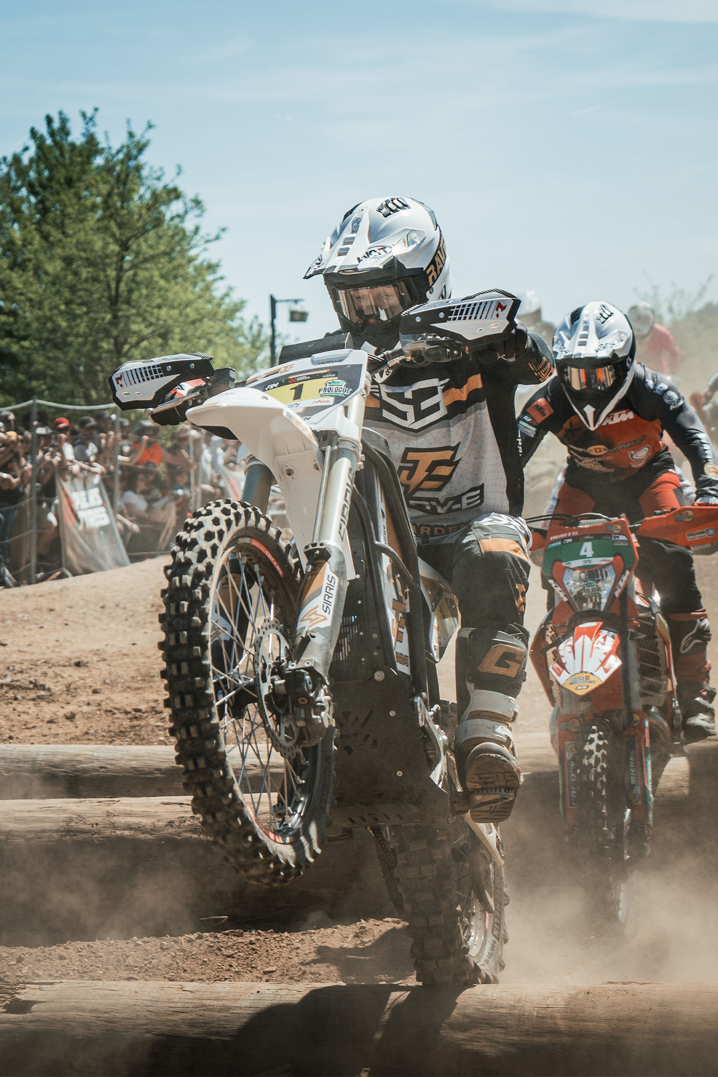 Motorcyclist in black gear jumping a dirt trail with bare trees and mountains in the background.