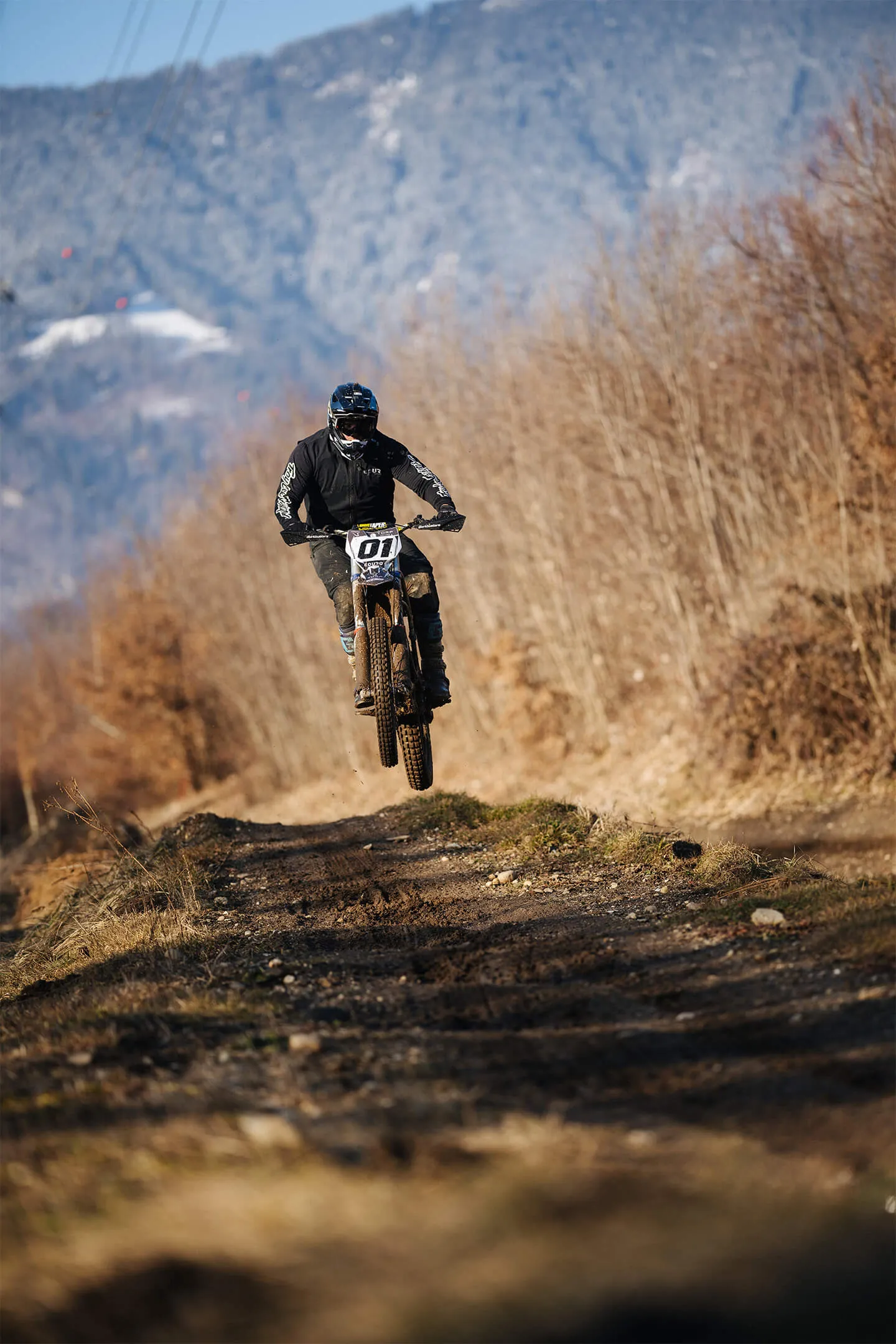 Motorcyclist in black gear jumping a dirt trail with bare trees and mountains in the background.