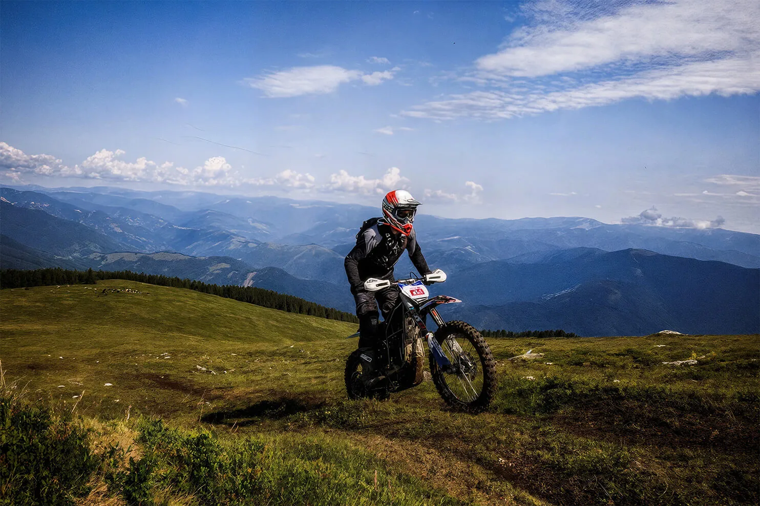 Motorcyclist wearing safety gear riding on a grassy mountain trail with a backdrop of rolling blue mountains under a partly cloudy sky.