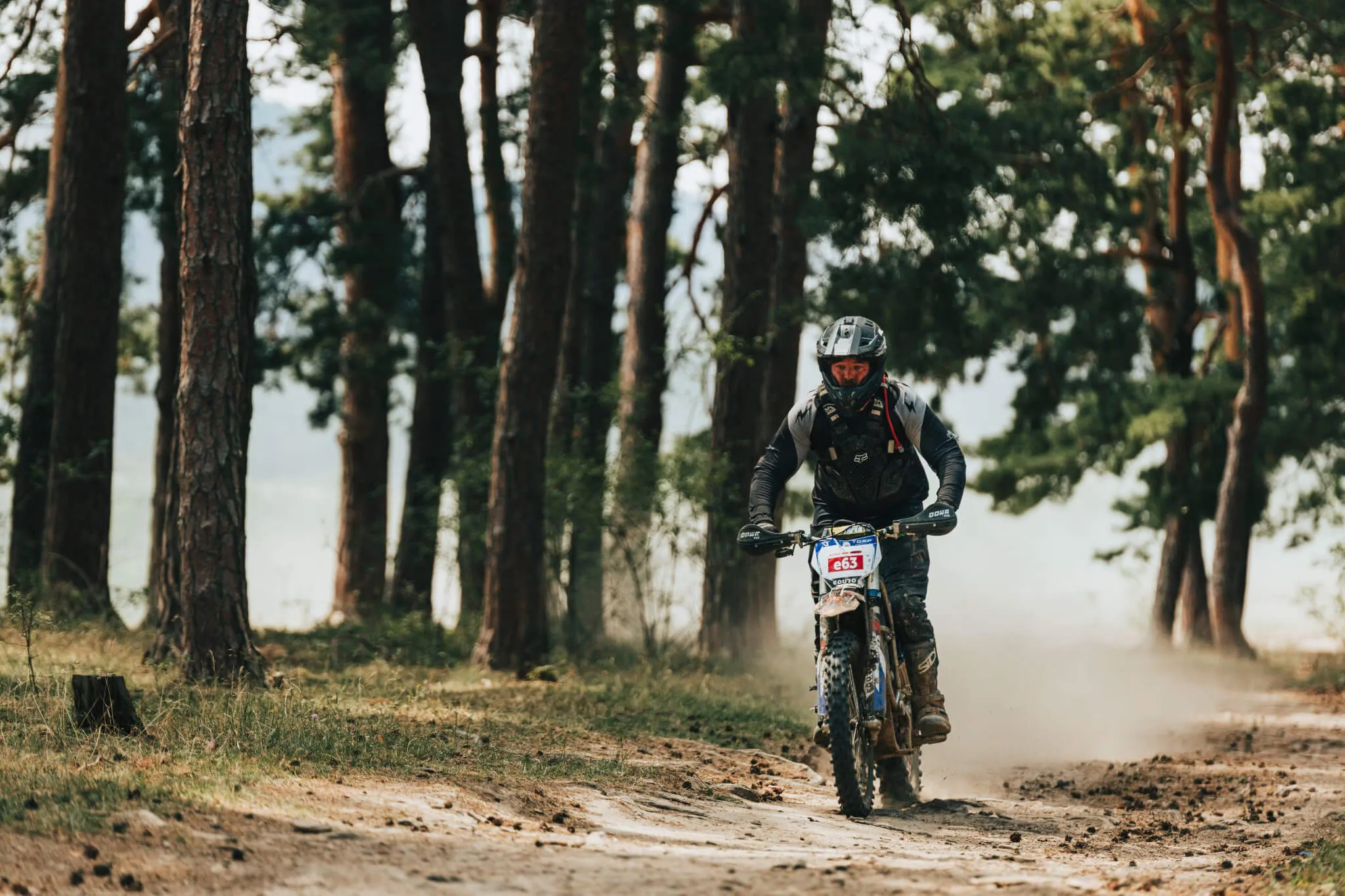 Motorcyclist wearing protective gear riding a dirt bike through a forest trail with dust rising behind.