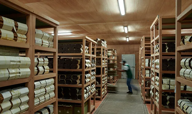 Warehouse aisles with wooden shelves stacked with rolled materials, and a worker in a green vest handling items on a trolley.