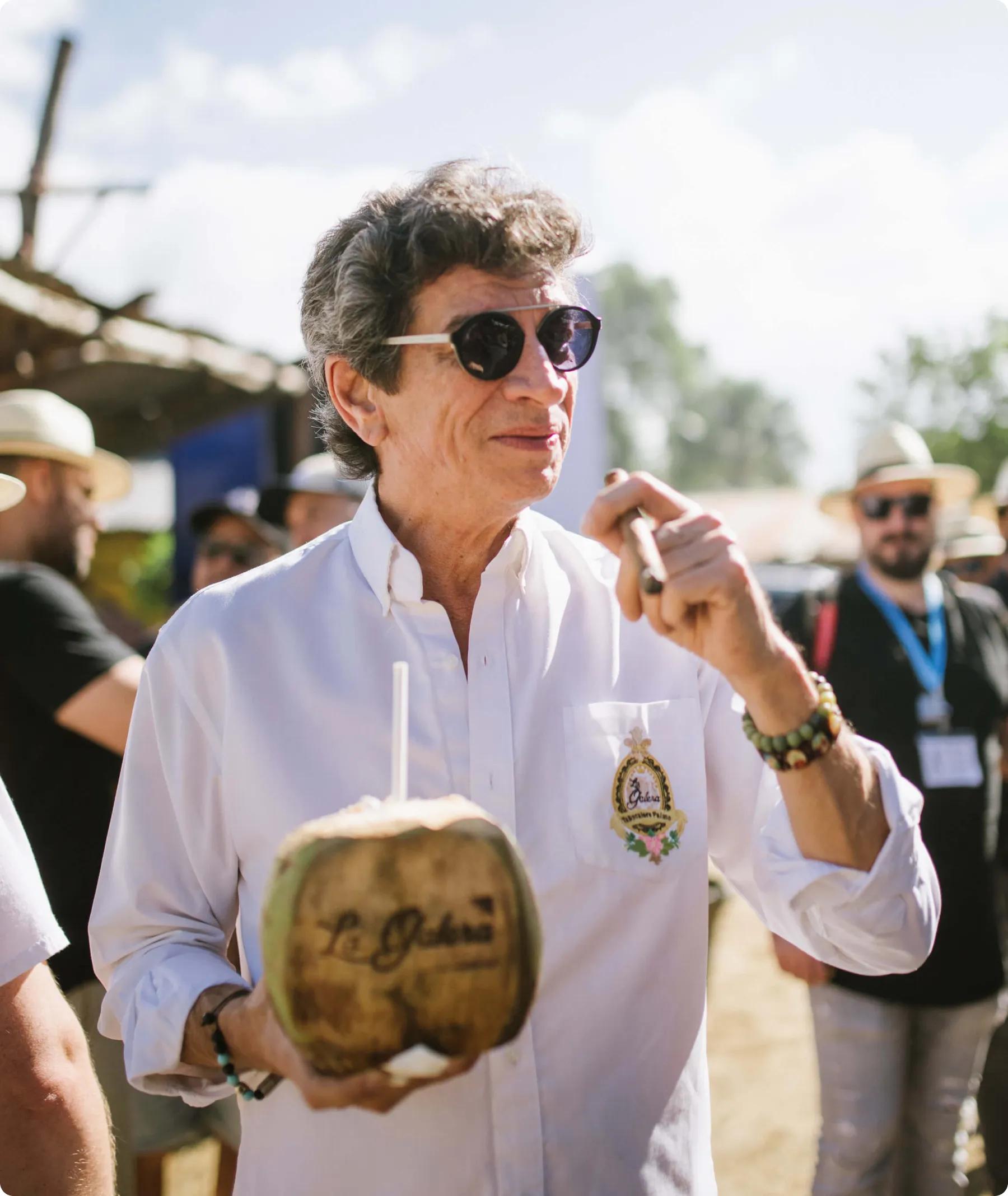 Man wearing sunglasses and a white shirt holding a coconut drink with a straw and a cigar outdoors.