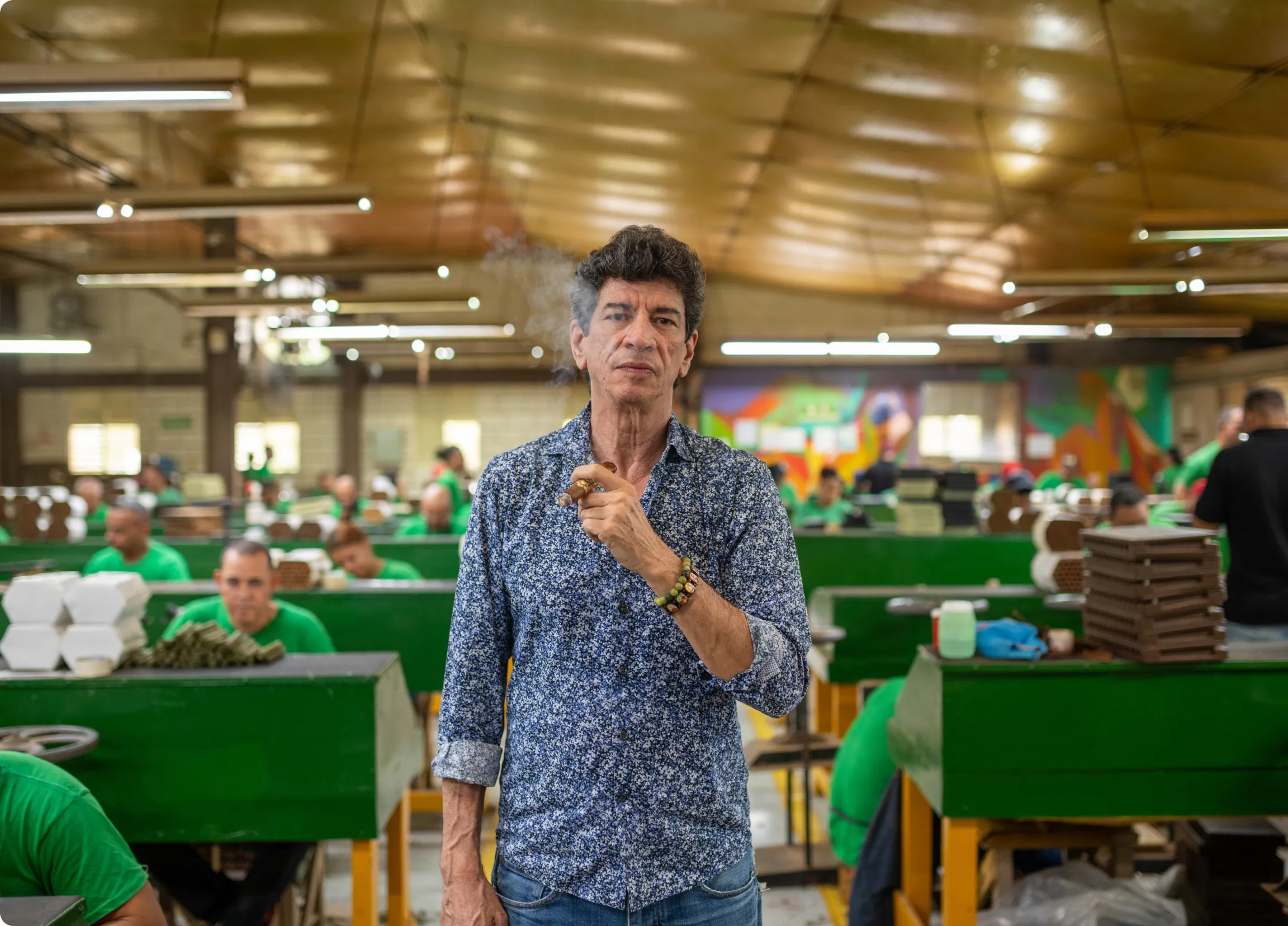 Man smoking a cigar standing in a factory with workers in green shirts processing cigars in the background.