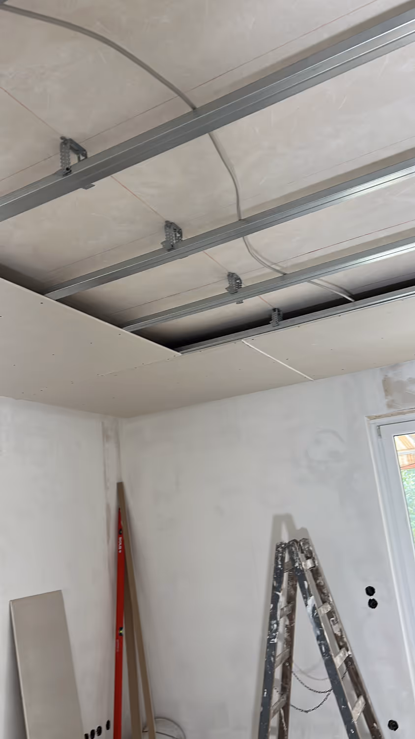 Raum im Bau mit freiliegenden Deckenmetallträgern, teilweise installierter Gipskartondecke, einer Leiter und Werkzeugen an unfertigen weißen Wänden.