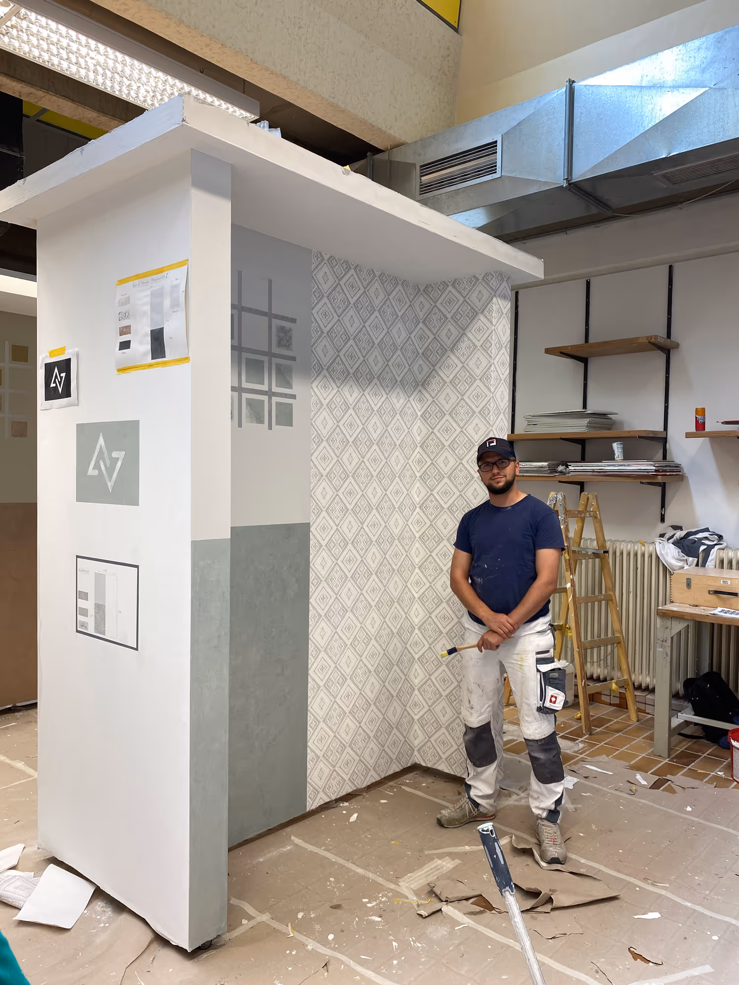 Maler steht in einer Werkstatt neben einer weiß-grau teilweise gestrichenen Wand mit einem Abschnitt aus diamantgemusterter Tapete.