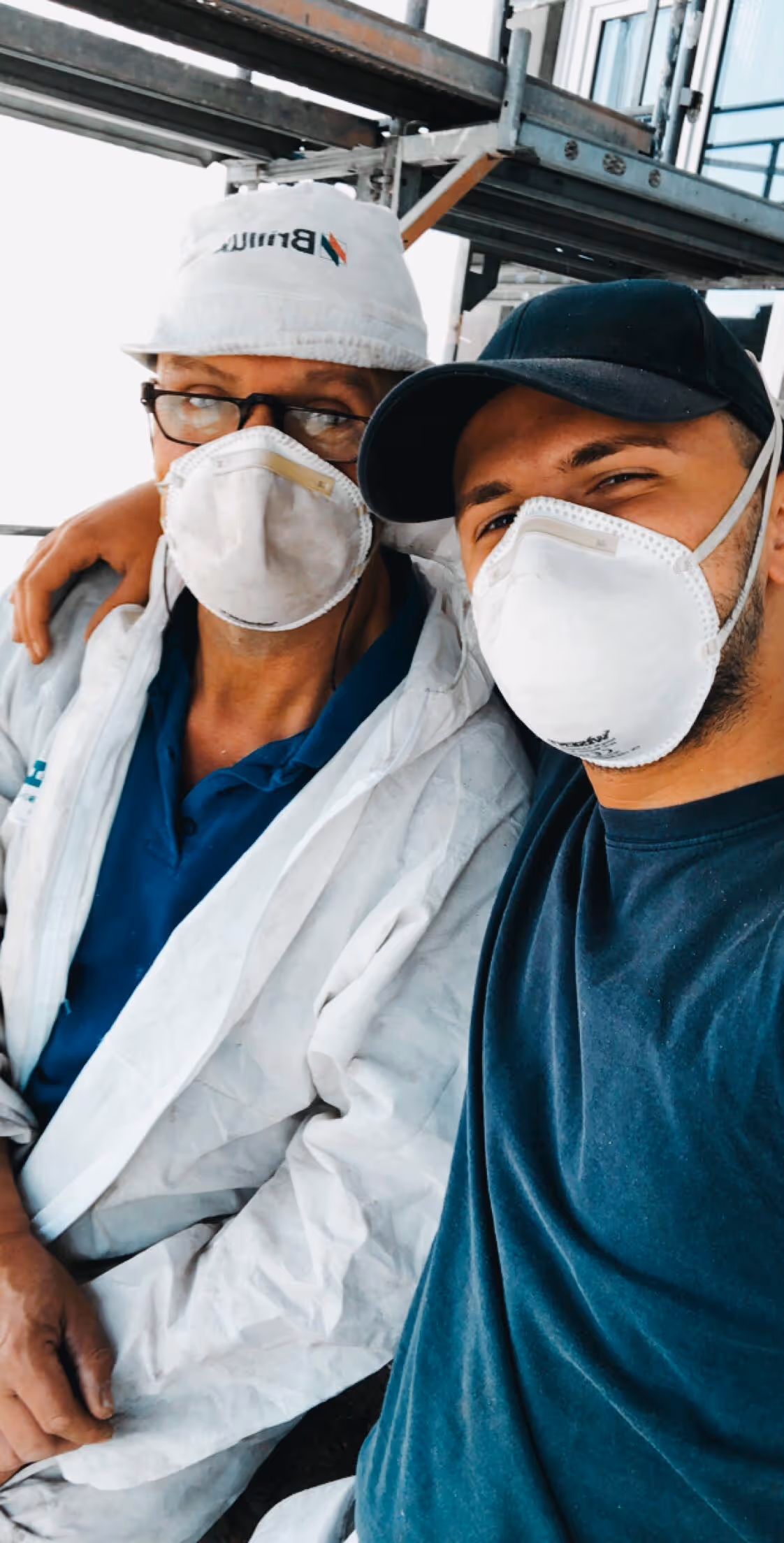 Zwei Männer mit Schutzmasken, einer in einem weißen Schutzanzug und Hut, der andere mit schwarzer Kappe und dunklem Shirt, sitzen dicht beieinander.