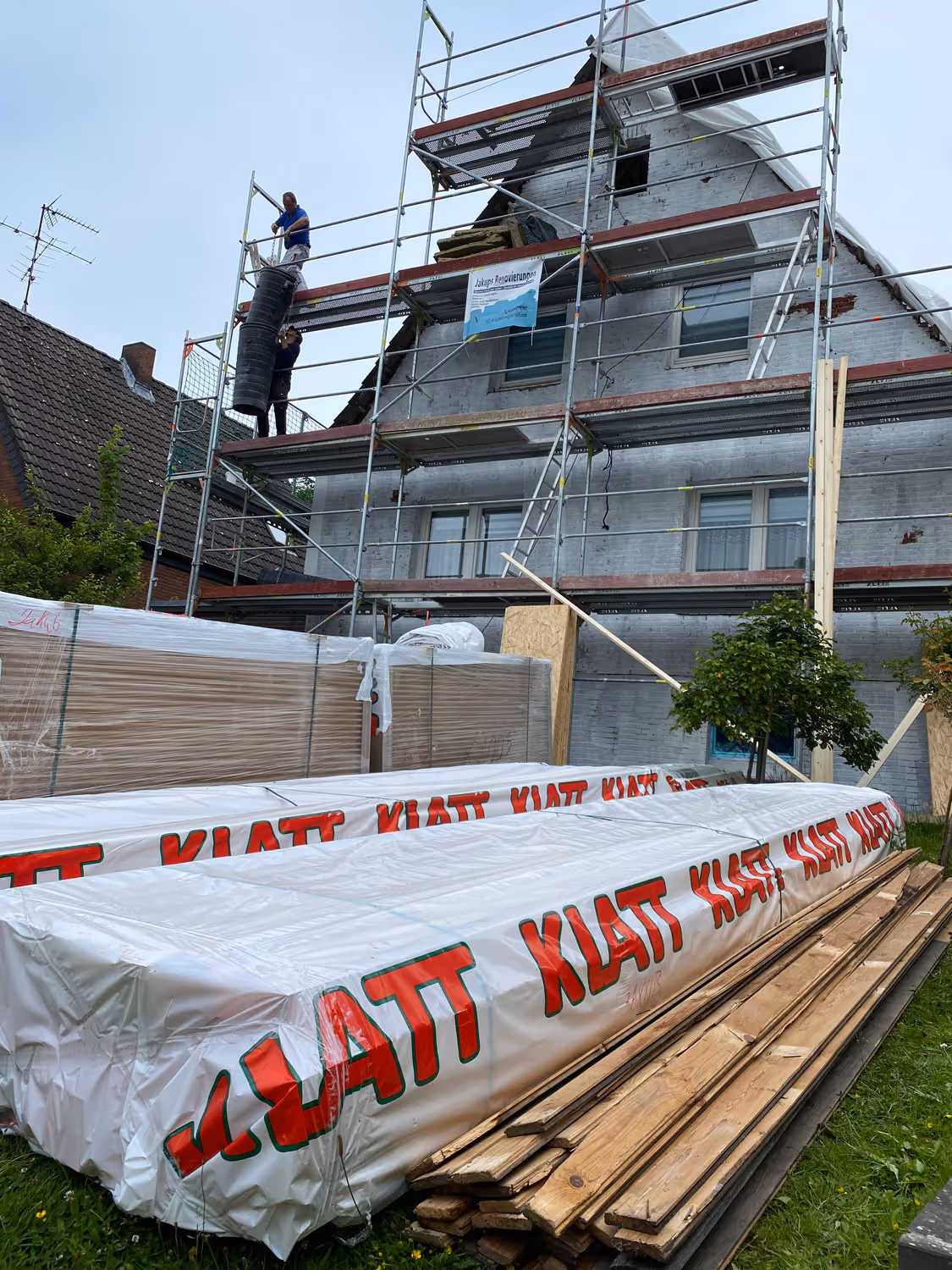 Zwei Bauarbeiter auf einem Gerüst entladen Materialien aus einer Rutsche neben einem Haus, das gerade renoviert wird.