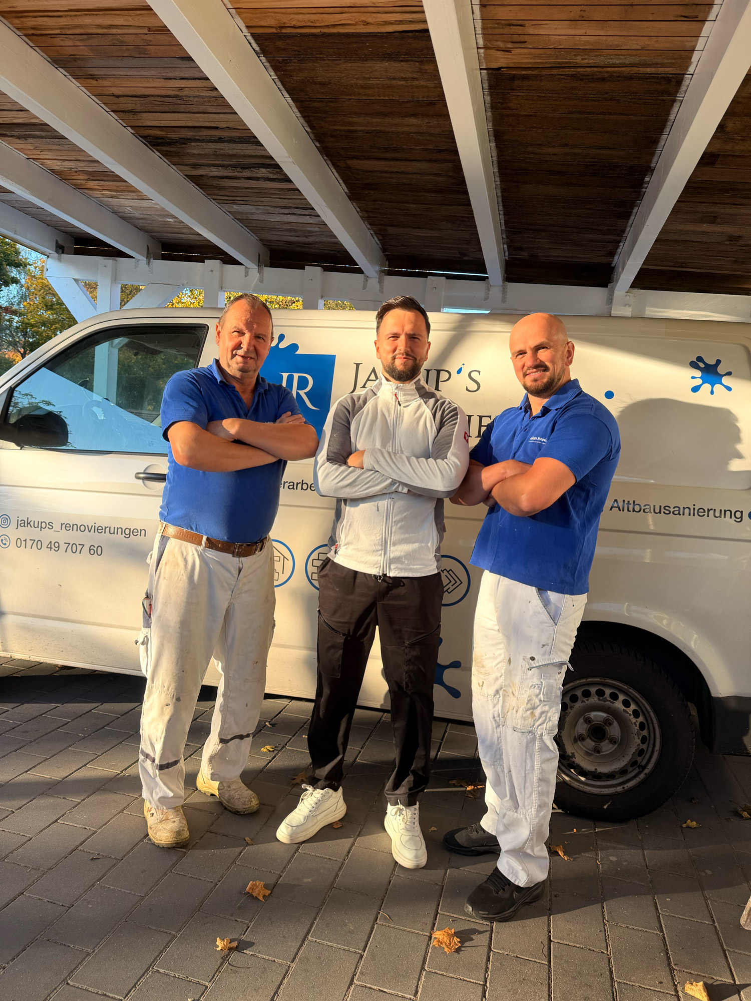 Drei Männer stehen mit verschränkten Armen vor einem weißen Arbeitswagen unter einem Holzdach.