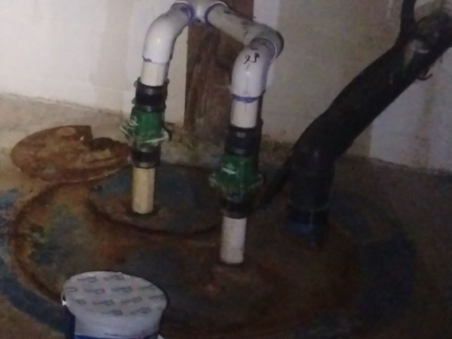 Rusted sump basin lid in a basement floor, two white discharge pipes with green check valves rising to the block wall